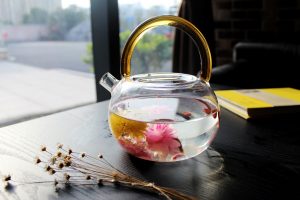 A clear glass teapot with a golden handle contains water and floating pink and yellow flowers, perfect for steeping holy cow tea. It sits on a dark table beside dried flowers and a closed yellow book, sunlight streaming through the window.