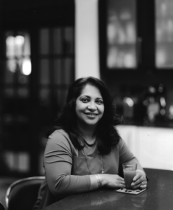 A woman with long dark hair, wearing a long-sleeved top and a necklace, sits at a table holding a glass of what might be Holy Cow Tea and smiling. The background shows shelves and cupboards, slightly out of focus in black and white.