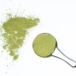 A metal scoop filled with Matcha - Ceremonial Grade rests on a white surface, with extra vibrant green powder scattered nearby.