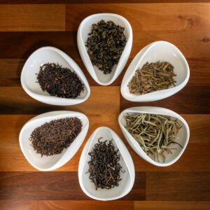 Six white bowls filled with assorted loose leaf teas are artfully arranged in a circle on a wooden surface, inviting you to embark on “A journey to discover what is Tea?”—a masterclass experience with a Tea Sommelier.