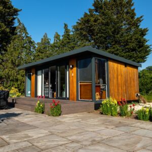 Immerse yourself in “A journey to discover what is Tea? Masterclass/Experience with Tea Sommelier” in a serene, contemporary wooden house featuring expansive glass windows atop a stone patio, surrounded by vibrant blooms and lush, towering trees under a brilliant blue sky.