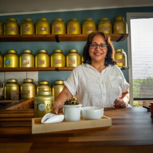 260527_Holy Cow-44 A woman in a white shirt stands behind a wooden counter with tea canisters in the background, leading “A journey to discover what is Tea? Masterclass/Experience with Tea Sommelier,” while a tray holding a teapot, cup, and loose tea leaves is displayed in the foreground.