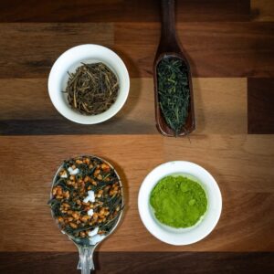 Experience the Green Tea Masterclass with a Tea Sommelier, featuring four varieties: loose leaf green tea in two bowls, powdered green tea in a small bowl, and a green tea blend with toasted rice in a spoon—all beautifully arranged on a wooden surface.