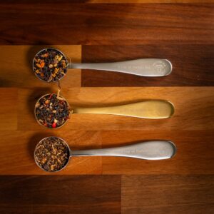 Three metal measuring spoons, filled with assorted loose leaf teas from the Chai Blends Masterclass/Experience with Tea Sommelier, are arranged horizontally on a wooden surface. The spoons showcase two silver and one gold hue.
