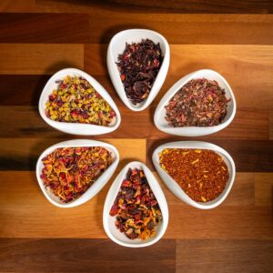 Six white bowls filled with a vibrant assortment of dried herbs and spices are artistically arranged in a flower-like circle on a wooden surface—an inviting presentation from the Holy Cow Tisanes Masterclass/Experience with Tea Sommelier.