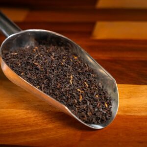 A metal scoop filled with loose Black Tea Masterclass/Experience with Tea Sommelier leaves rests on a wooden surface with alternating light and dark brown stripes.