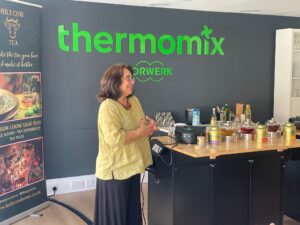 A woman stands and smiles whilst speaking next to a kitchen worktop with tea jars and teapots. Behind her are "thermomix" and "Vorwerk" signs on the wall, and a banner for Holy Cow Tea.
