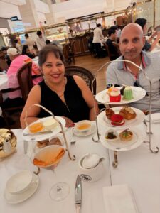 A smiling couple sits at a restaurant table set for afternoon tea, with a three-tiered tray of colourful pastries and snacks, cups of tea, and a cocktail. Other diners are seated at tables in the background.