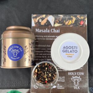 A tin labelled "Holy Cow Tea Masala Chai," a cup labelled "Agosti Gelato Masala Chai," loose tea leaves in a glass container, and a menu card with Masala Chai information are arranged on a table.