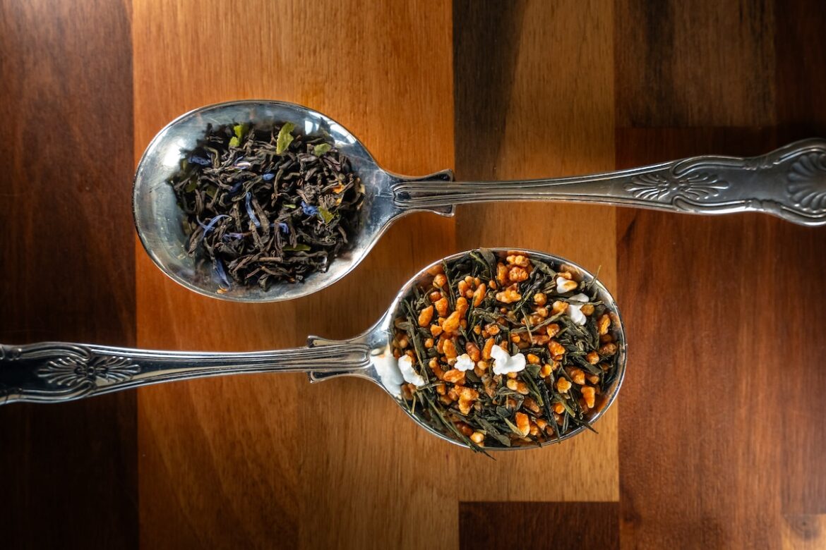 Two ornate silver spoons on a wooden surface, one holding loose black tea leaves and the other holding a colourful blend of green tea, puffed rice, and white bits, possibly Genmaicha with popcorn.