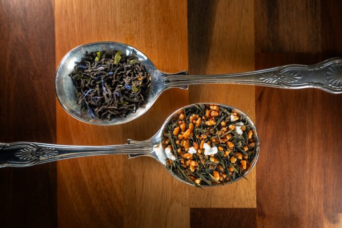 Two ornate silver spoons on a wooden surface, one holding loose black tea leaves and the other holding a colourful blend of green tea, puffed rice, and white bits, possibly Genmaicha with popcorn.