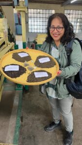 A smiling woman holds a round tray displaying four types of tea leaves labelled: WHOLE LEAF, DUST, FANINGS, and BROKEN, inside what appears to be a tea processing factory.