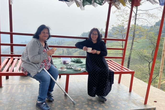Two women sit on a red bench under a canopy, enjoying drinks with snacks on a table. One woman has a crutch. Lush greenery and mountains are visible in the misty background.