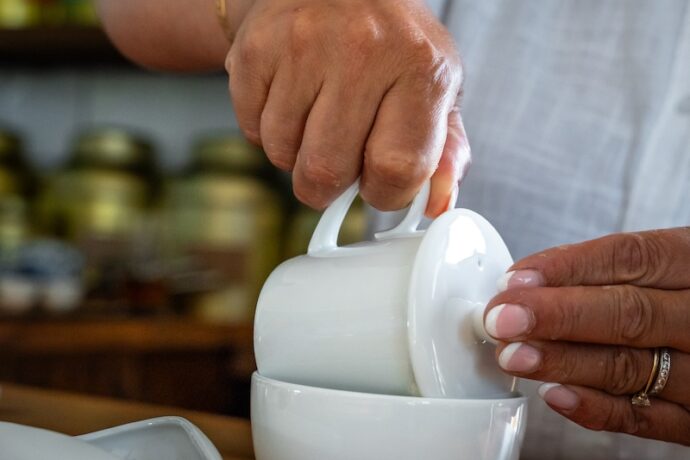 A person in a white shirt lifts the lid off a white bowl on a wooden table, with a small white mortar and pestle nearby and jars on shelves in the blurry background.