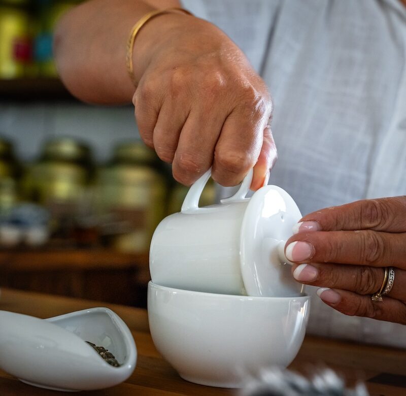 A person in a white shirt lifts the lid off a white bowl on a wooden table, with a small white mortar and pestle nearby and jars on shelves in the blurry background.