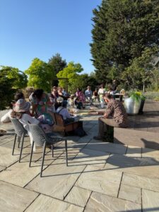 A group of people sit outdoors on a sunny day, listening to a speaker near a wooden decking, surrounded by trees and greenery. Some people are seated on chairs, others on the ground.