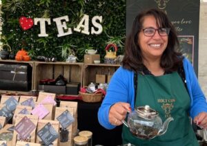 A smiling woman in glasses and a green apron pours tea at a market stall with tea packets and a “TEAS” sign behind her. The stall has teapots and green foliage decor.