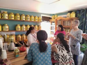 Five women gather and chat in a cosy tea room with wooden shelves lined with gold canisters. Some hold teacups while smiling and engaging in conversation. Sunlight brightens the room, creating a warm, friendly atmosphere.